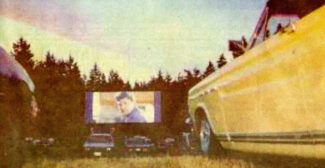 View of a Drive-In movie screen with a sleek yellow car to the right some distance away from the movie