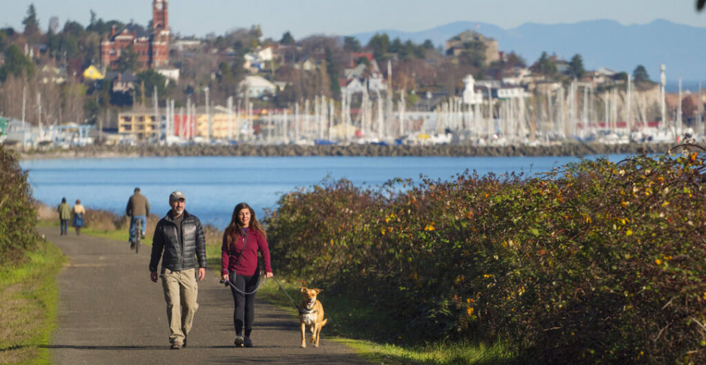 A man and woman walk their dog on the Larry Scott Trail along the water with Port Townsend's homes and buildings in the background