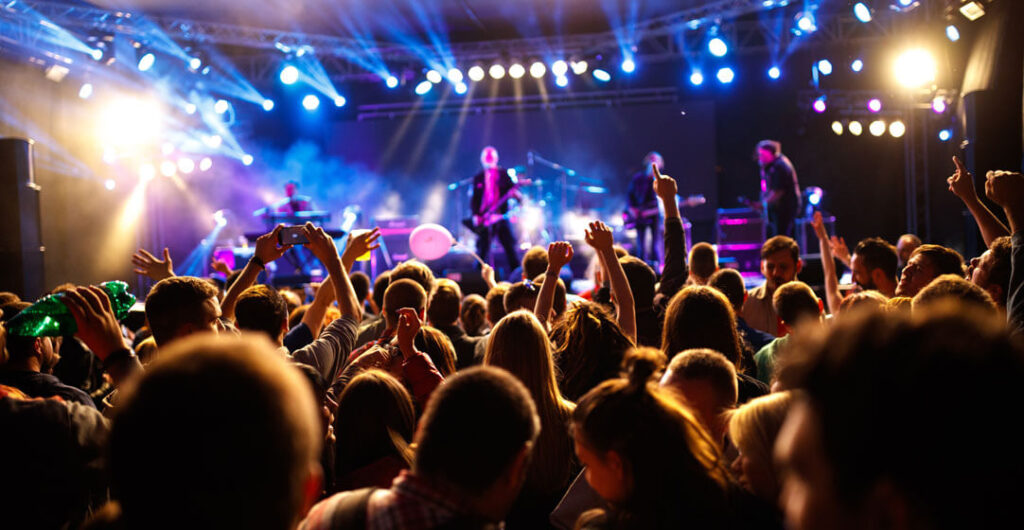 A crowd listens to a rock concert under the lights of a stage