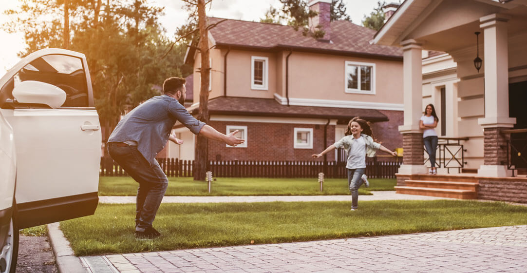 A young girl runs into the arms of her father in the front yard, to illustrate the importance of umbrella insurance.
