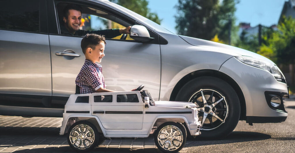A man is parked in his driveway in a silver SUV, and his son is beside him in silver toy car, to illustrate the importance of umbrella  insurance. 