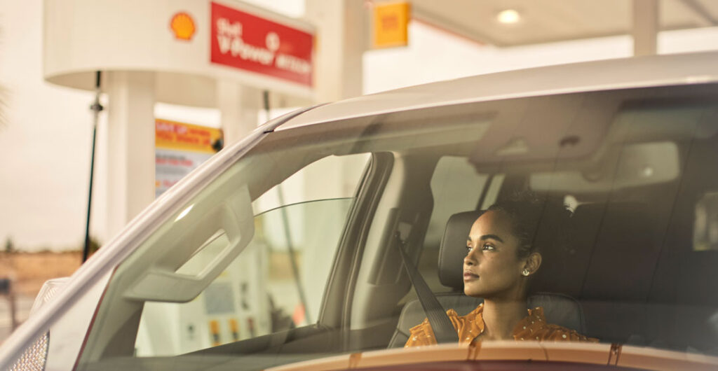 A female passenger in a car looks out the window, the car is parked at a Shell gas station, to illustrate that you can get AAA members can qualify for a gas discount at Shell stations.